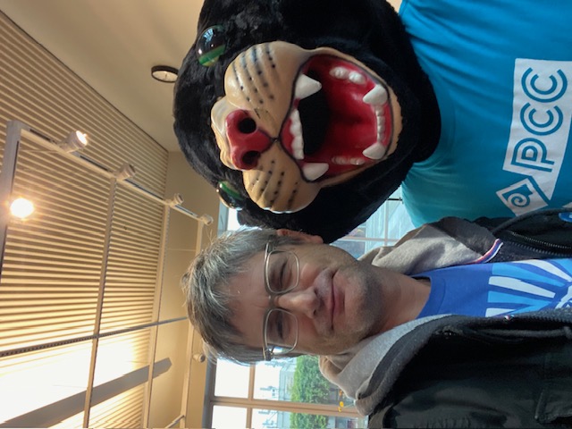 A photo of Dan Davis wearing glasses, with a blue ffap t-shirt, black hooded jacket. Also in the photo is Pinky the Panther wearing a blue PCC t-shirt.