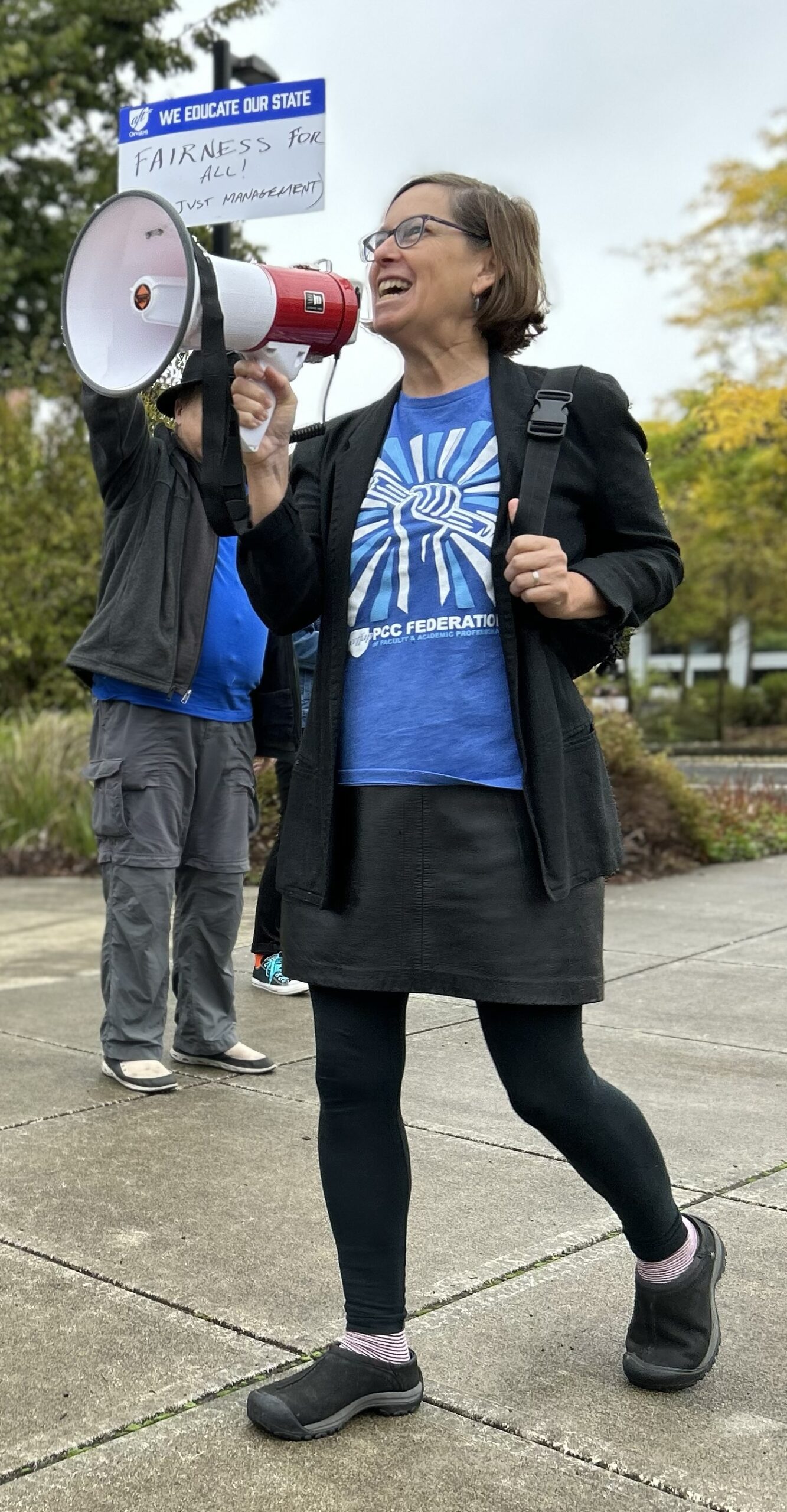 Photo of Julie Hastings carrying a bullhorn, wearing an ffap blue shirt, black skirt, and black leggings. A person in the background is holding an FFAP sign that is not readable.