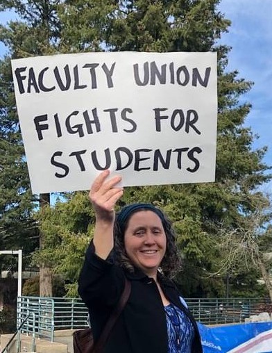A photo of Marianne Stupfel-Wallace holding a sign that reads Faculty Union Fights for Students. Wearing a black jacket with a blue FFAP button