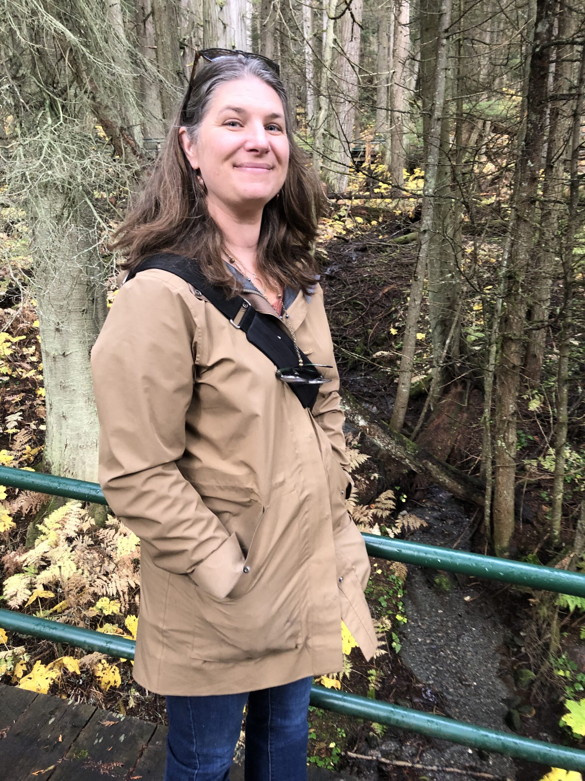 A photo of Nichole Reding standing on a bridge in a forest. Wearing a light brown jacket with a cross body bag and jeans. She also has sunglasses on her head.