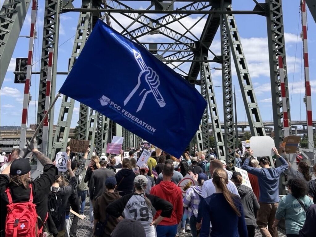 Photograph of a crowd of people, viewed from behind, walking across a bridge. One figure wearing a red backpack and holding up a large blue flag with a white line drawing of a fist holding a pencil with text below the drawing reading: 'PCC Federation of Faculty and Academic Professionals'